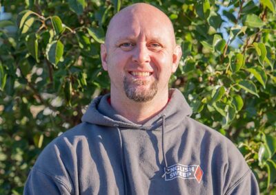 Greg S. - Sales & Service Tech has a shaved head with a goatee. He is wearing an English Sweep gray hooded pullover with greenery in the background.