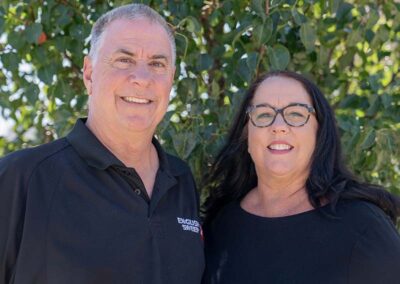 Gregg and Cami owners, both wearing black shirts Greg's says English Sweep. He has short gray hair blue eyes and a nice smiles. Cami has long black hair, beautiful smile and is wearing glasses. In the background are green bushes.