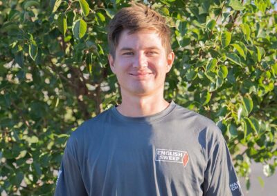 Kylan B. - Project Coach has short red hair with a nice smile. He is wearing a long sleeve English Sweep T in gray with greenery in the background.
