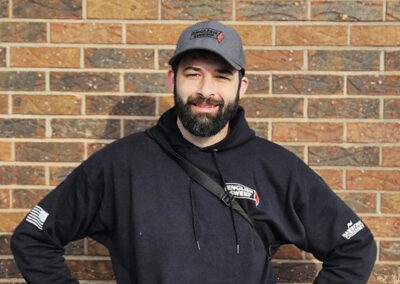 Kyle G. is wearing an English Sweep ball cap. He has short black hair, mustache, beard and brows. He is wearing a black hooded pullover with a brick wall in the background.