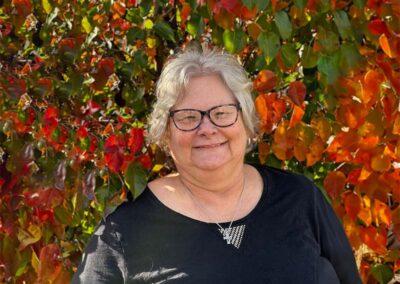 Lorelei K. is a Customer Service Rep with short gray hair parted in the middle wearing glasses with a nice smile. She. is wearing a black blouse with a necklace and has autumn colored bushes in the background.