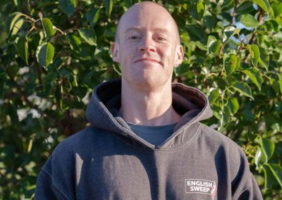 Tyler B. - Project Coach appears to be standing at attention has a bald head and reddish brown eye brows. He is wearing a hooded English Sweep pullover in dark gray and their is greenery in the background.
