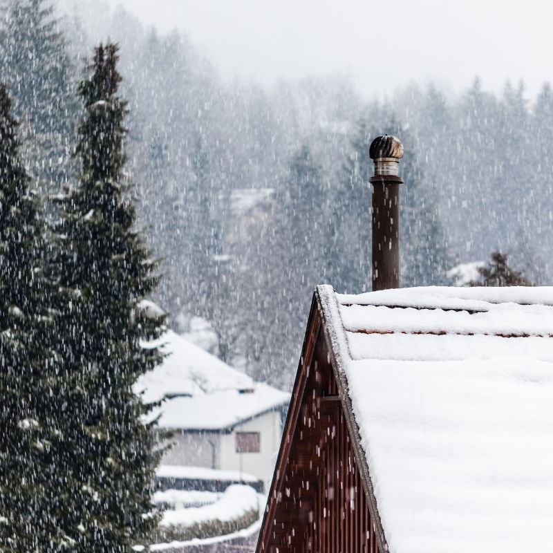 a snowy rooftop with a chimney pipe coming out of it and a snowy landscape with trees behind