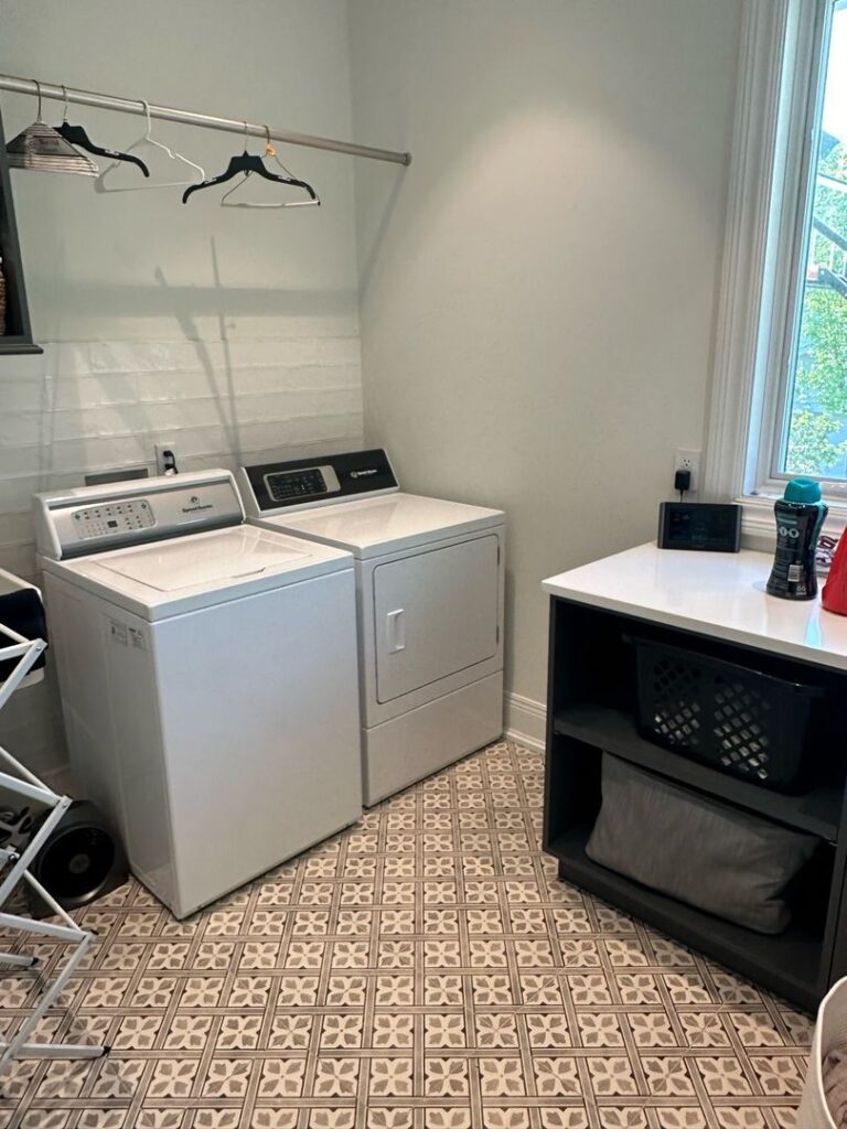 A tidy laundry room with a side-by-side washer and dryer, shelving unit, and patterned tile flooring.
