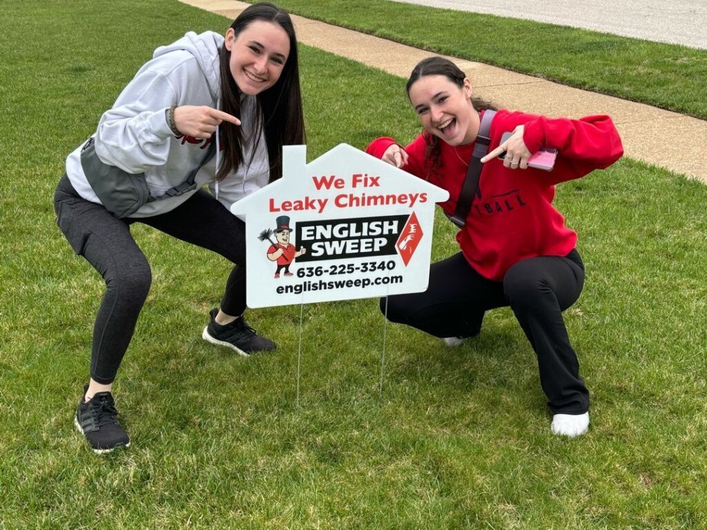 Two people crouching on grass, pointing at an English Sweep yard sign for chimney repair services.